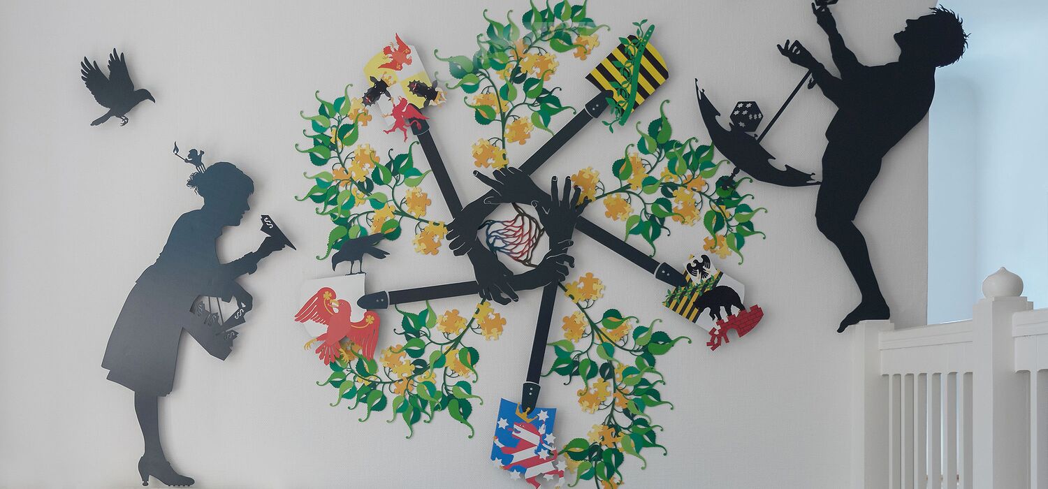 Ländernotarkasse Leipzig, Projekt Spectrum freies Notariat von Wolf v. Waldow – Treppenhauswand mit Emblem aus farbig bemaltem, Stahlblech mit Wappen der 5 Neuen Bundesländer und Pflanzenzweigen; 2 Figuren die Aufgaben der Kasse zeigen