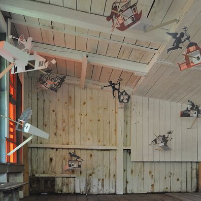 innenansicht Projektraum "Hühnerhaus Volksdorf" mit Installation Neue Heimat von Wolf v. Waldow: Holzschuppen mit 9 Objekten in Form länglicher Überwachungs-Kameras, deren Fronten mit allegorischen Motiven aus laminiertem, schwarzem Papier verkleidet sind