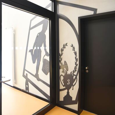 Ausgangstür UG Sporthalle Kniprodestr. Projekt Hlifestellung – scherenschnittartige Figur auf Wand von Wolf v. Waldow, Motiv: Sportler macht Dehnübung und gießt mit Gießkanne Pokal für 3. Platz, aus dem Lorbeerkranz wächst.