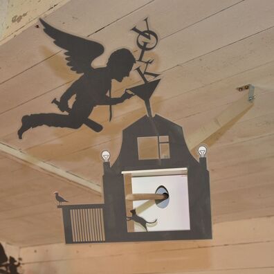 Projektraum "Hühnerhaus Volksdorf" mit Installation Neue Heimat von Wolf v. Waldow: Objekt in Form länglicher Überwachungs-Kamera, Front mit Motiv aus laminierten Papier: Engel mit Basballschläger, der durch Trichter sas Wort "Volk" in Haus einfüllt.