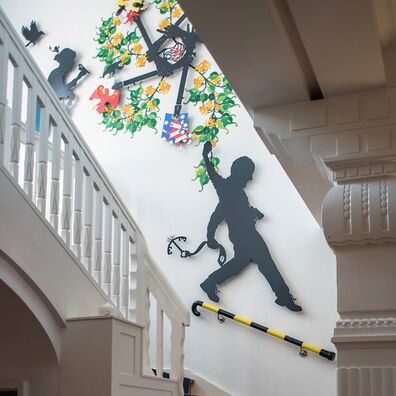 Ländernotarkasse Leipzig, Projekt Spectrum freies Notariat von Wolf v. Waldow – Treppenhauswand mit Emblem aus farbig bemaltem, gelasertem Stahlblech von Unten; scherenschnittartige Figur herabsteigend über dem wie Krückstock geformten Geländer.