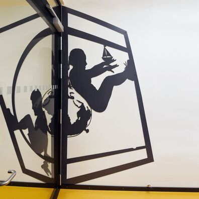 Eingangstür UG Sporthalle Kniprodestr. Projekt Hlifestellung – 2 scherenschnittartige Figuren auf Wand von Wolf v. Waldow, Motiv: Sportlerin u. Sportler lehnen sich Rücken an Rücken gegen Medizinball, der die Form einer deformierten Weltkugel hat.