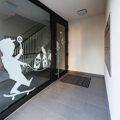 Eingangstür und Glasfenster mit weiß aufgedrucktem Silhouetten-Motiv von Wolf v. Waldow: Kleiner Junge auf Skateboard balanciert Spielzeugbahn aus verschidenen, symbolischen Hauszeichen.