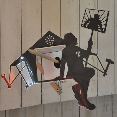 Projektraum "Hühnerhaus Volksdorf" mit Installation Neue Heimat von Wolf v. Waldow: Objekt in Form länglicher Überwachungs-Kamera, Front mit Motiv aus laminierten Papier: Mann mit signet "Arbeitsamt" sitzt vor Zelt und hält Schild mit Politiker hoch.