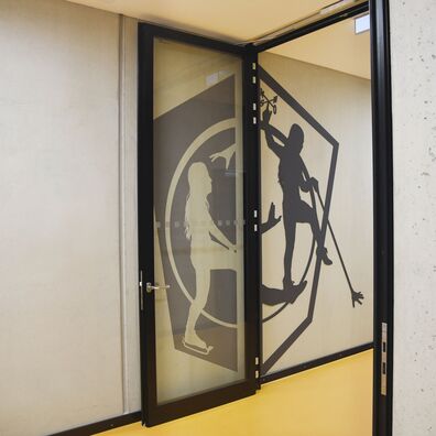 Rauchfangtür offen Sporthalle Kniprodestr. Projekt Hlifestellung – 2 scherenschnittartige Figuren (1. Stahl, 2. gemalt) auf Wand von Wolf v. Waldow, Motiv: Sportlerin balanciert auf Fuß der anderen, die ihr zur Unterstützung die Hand reicht.
