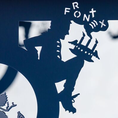 Tisch aus schwarz gepulvertem Stahl von Wolf v. Waldow; scherenschnittartige Figur sitzt zwischen Zarge und Tischbein, Motiv: Mann mit Geburtstagstorte auf Knie; aufsteifender Rauch formt das Wort "Frontex"
