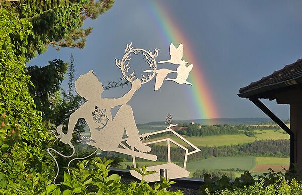Blick auf eine figürliche Allegorie (Silhouette) zum Thema Klimawandel in einem Garten mit Regenbogen im Hintergrund