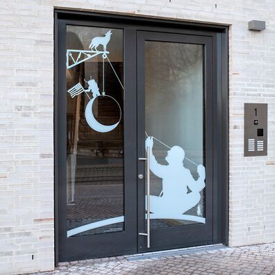 Eingangstür und Glasfenster mit weiß aufgedrucktem Silhouetten-Motiv von Wolf v. Waldow: Mann zieht Mond an Flaschenzug hoch, dessen Rolle über ein auskragendes Gasthaus-Schild mit heulendem Wolf läuft.