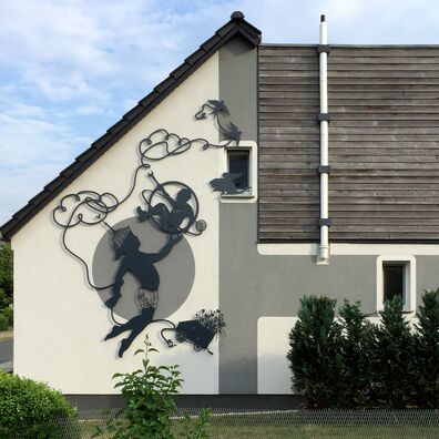 Ansicht Hausfassade in Goyatz, Spreewald mit Silhouetten-Arbeit von Wolf v. Waldow; Motiv: Springender Junge, der ein Zahnrad in der Hand hält in dem ein Mann sitzt, der nach einem Seil greift, das sich wolkenartig über ihm kringelt.