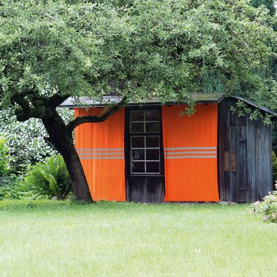Ansicht Projektraum "Hühnerhaus Volksdorf" mit Installation Neue Heimat von Wolf v. Waldow: Holzschuppen mit Verkleidung aus Orange-farbigen Stoffbahnen mit Reflektorstreifen im Garten unter Baum.