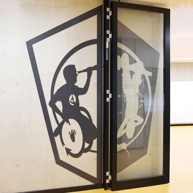 Rauchfangtür offen Sporthalle Kniprodestr. Projekt Hlifestellung – 2 scherenschnittartige Figuren (1. Stahl, 2. gemalt) auf Wand von Wolf v. Waldow, Motiv: Sportler im Rollstuhl hält Fernrohr an dem anderer Sportler kopfüber hängt (Schweinebaumeln).