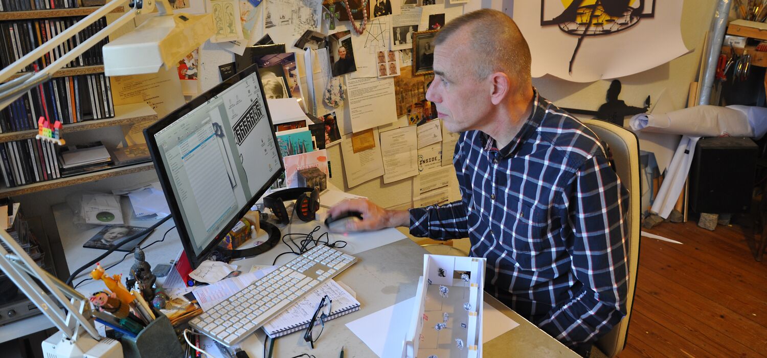 Wolf v. Waldow im Atelier sitzt am Computer; hinter ihm an der Wand Zeichnungen, im Vordergrund Arbeitsmodell für Ausstellung