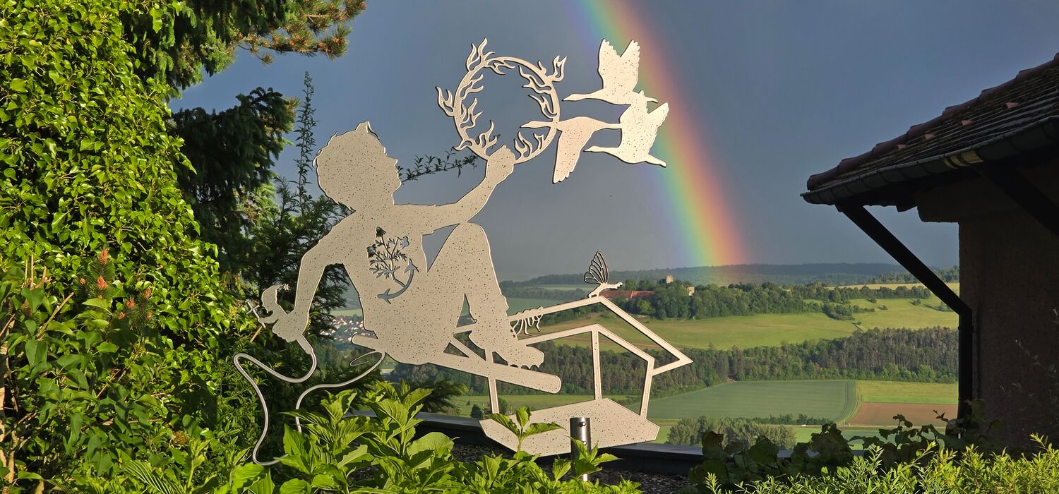 Allegorische Figur, aus Aluminium als Silhouette (Scherenschnitt) auf einer hohen Stange vor grünen Sträuchern; Regenbogen; Motiv: Junge sitzt auf einer Art Vogelhäusschen und hält einen brenenden Reifen durch den Gänse fliegen.