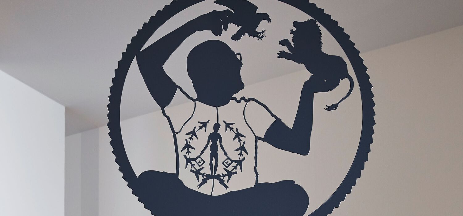 Scherenschnittartige, allegorische Figur in rundem Rahmen von Wolf v. Waldow, Motiv: Mann hält angreifenden Adler und kämpfenden Löwen in Händen gegenüber; auf Shirt Figur in "Peace"-Haltung, umgeben von Flugzeugen