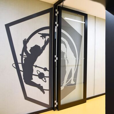 Rauchfangtür offen Sporthalle Kniprodestr. Projekt Hlifestellung – 2 scherenschnittartige Figuren (1. Stahl, 2. gemalt) auf Wand von Wolf v. Waldow, Motiv: Sportlerin springt in einen Planetenring, andere Sportlerin holt Schwung und weist ihr die Richtung