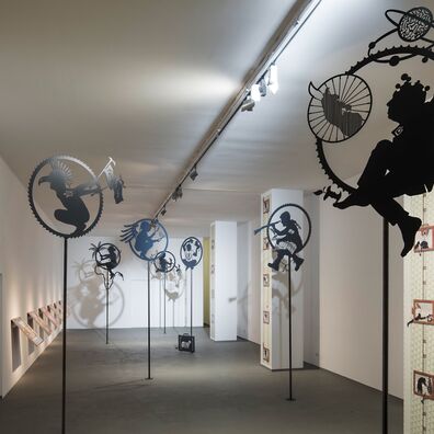 Blick in Solo-Ausstellung "Die Hand vor Augen" von Wolf v. Waldow im "Neuen Kunstverein Wuppertal" mit 7 scherenschnittartigen, figürl. Emblemen auf Stangen; Tapetenarbeit "Refugee camp" an 3 Pfeilern, die  Raum gliedern; Arbeit "Competitions" auf Haltern