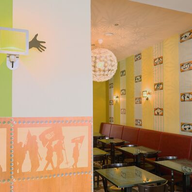 Blick in den hinteren Gastraum des Café Bierhimmel Berlin; Wandflächen gelb abwechselnd mit einzelnen Tapetenstreifen (Tapete "Traveller" von Wolf v. Waldow); rote Polsterbänke, Cafétischchen mit Glasplatten
