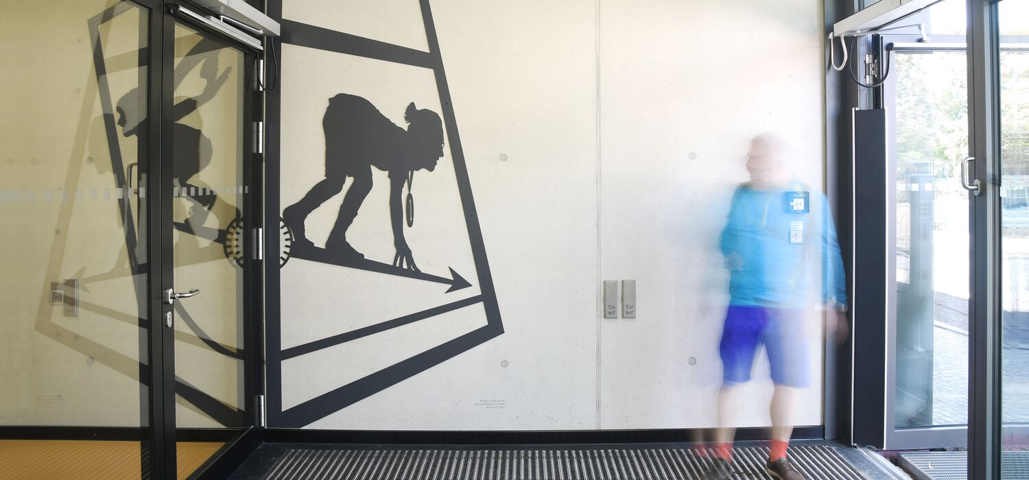 Foyer Sporthalle Kniprodestr. Projekt Hlifestellung – scherenschnittartige Figur aus Stahl auf Wand von Wolf v. Waldow, Motiv: Sportler in Startposition für Sprint, gegeüber sSportler in Startposition losfliegen.