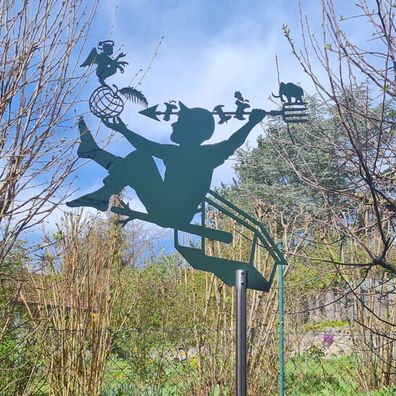 Allegorische Figur, aus Aluminium als Silhouette (Scherenschnitt) auf einer hohen Stange vor kahlen Sträuchern; Motiv: Junge sitzt auf einer Art Vogelhäusschen und zielt mit Pfeil auf Erdkugel-Apfel auf dem ein Engel sitzt.