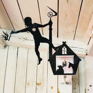 Projektraum "Hühnerhaus Volksdorf" mit Installation Neue Heimat von Wolf v. Waldow: Objekt in Form länglicher Überwachungs-Kamera, Front mit Motiv aus laminierten Papier das Frau, die auf Krippe mit Minarett balanciert und brennendes Streichholz hält.