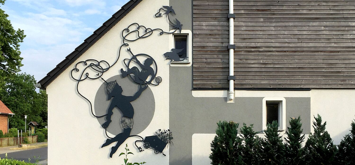 Ansicht Hausfassade in Goyatz, Spreewald mit Silhouetten-Arbeit von Wolf v. Waldow; Motiv: Springender Junge, der ein Zahnrad in der Hand hält in dem ein Mann sitzt, der nach einem Seil greift, das sich wolkenartig über ihm kringelt.