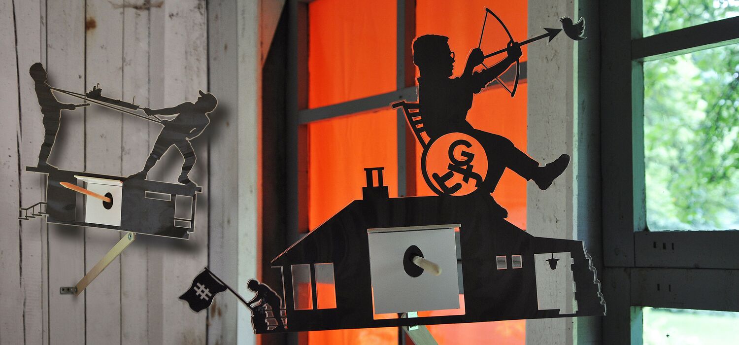 Projektraum "Hühnerhaus Volksdorf" mit Installation Neue Heimat von Wolf v. Waldow: Objekt in Form länglicher Überwachungs-Kamera, Front mit Motiv aus laminierten Papier: Frau im Rollstuhl auf Hausdach zielt mit Pfeil auf Vogel