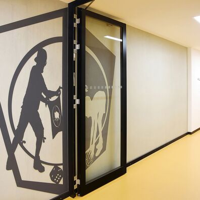 Rauchfangtür offen Sporthalle Kniprodestr. Projekt Hlifestellung – 2 scherenschnittartige Figuren (1. Stahl, 2. gemalt) auf Wand von Wolf v. Waldow, Motiv: Sportler hält Tuch mit Mammut als Ziel hoch, 2. Sportler bückt sich nach eckiger Form um zu werfen.