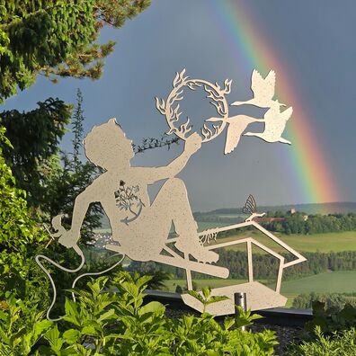 Allegorische Figur aus Aluminium als Silhouette (Scherenschnitt) auf einer hohen Stange vor Landschaft mit Regenbogen; Motiv: Junge sitzt auf einer Art Vogelhäusschen und hält einen brenenden Reifen durch den Gänse fliegen.