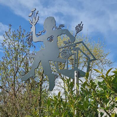 Allegorische Figur, aus Aluminium als Silhouette (Scherenschnitt) auf einer hohen Stange vor grünen Sträuchern; Motiv: Mädchen sitzt auf einer Art Vogelhäusschen und hält einen Löwenzahn in der einen und eine Rose in der anderen Hand.